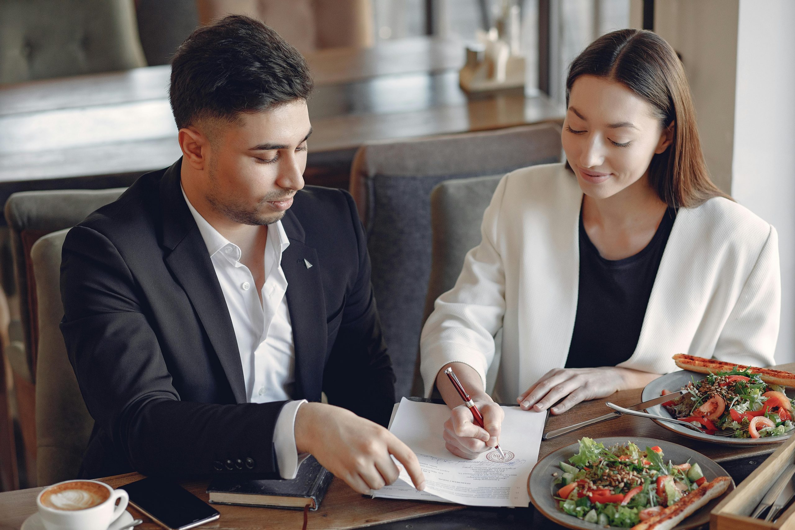 Concentrated multiethnic colleagues in formal clothes sitting at table in cafe with cup of espresso and plates of salad while signing documents