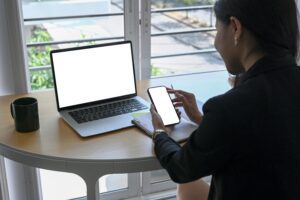 A lady looking at a cellphone at a desk with a laptop